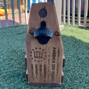 May include: A wooden bottle opener with a dark brown finish. It features a black metal opener that reads "OPEN HERE". The design includes the text "1776 2026" and "AMERICA 250 YEARS".