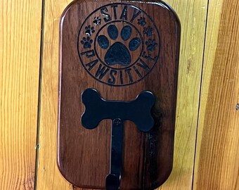 May include: A wooden dog leash holder with a dark stain. The holder features a black bone-shaped hook and a circular design with the words "STAY PAWSITIVE" and a paw print. The holder is mounted on a wooden wall.