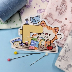 May include: A cute orange cat with a pink nose is sewing a colourful patchwork quilt on a yellow sewing machine. A blue bird is perched on the machine, and there are red and yellow pins scattered around.