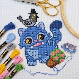Peut inclure: Une broderie au point de croix représentant un chat bleu aux grands yeux jaunes, un oiseau portant un chapeau et un pot de fleurs roses. Des fils à broder de différentes couleurs, des ciseaux dorés et un cercle à broder sont également visibles.