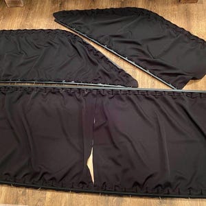 May include: Three black fabric window blinds with a pleated design. The blinds are lined up horizontally with the top blind slightly overlapping the middle blind. The bottom blind is the longest and has a decorative trim around the edges.