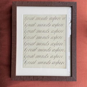Könnte beinhalten: Ein gerahmter Druck mit dem Satz „Great minds aspire“ in eleganter Schreibschrift. Der Druck ist in einem weißen Passepartout und einem dunkelbraunen Holzrahmen eingefasst, was eine klassische Ästhetik schafft. Der Hintergrund ist ein strukturiertes, cremefarbenes Papier.