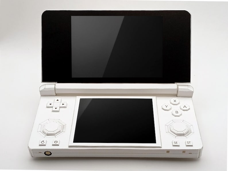 May include: A white handheld gaming console with two black screens, one on top and one below. The console has a directional pad, buttons, and a central control. The device is open, revealing the screens.
