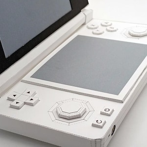 May include: A white handheld gaming console with a clamshell design. The console features a top screen, a bottom screen, and various buttons, including a directional pad, face buttons, and a central control stick. The device is made of white material.