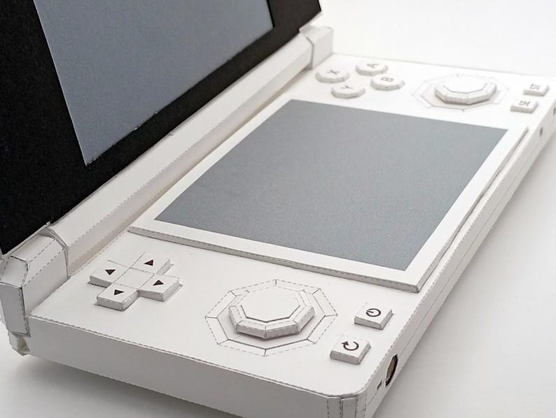 May include: A white handheld gaming console with a clamshell design. The console features a top screen, a bottom screen, and various buttons, including a directional pad, face buttons, and a central control stick. The device is made of white material.