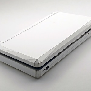 May include: A white, origami-style handheld gaming console, folded from paper. The device is shown partially open, revealing the interior. The design features sharp angles and a clean, minimalist aesthetic. The console is set against a plain white background.