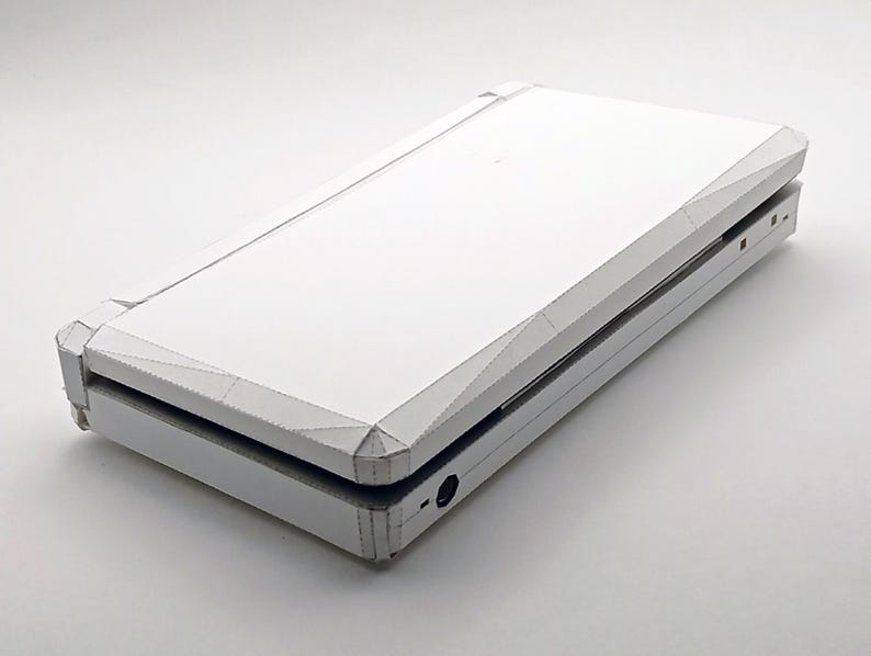 May include: A white, origami-style handheld gaming console, folded from paper. The device is shown partially open, revealing the interior. The design features sharp angles and a clean, minimalist aesthetic. The console is set against a plain white background.