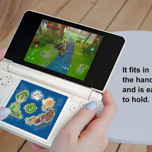 May include: A white handheld gaming console with two screens, one displaying a game scene and the other a map. The text "It fits in the hand and is easy to hold" is visible. The console has buttons and a directional pad.