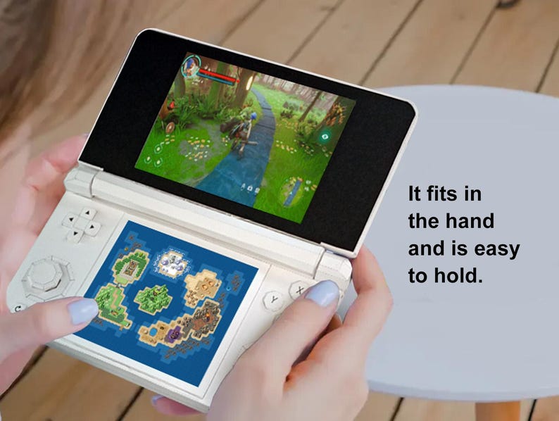 May include: A white handheld gaming console with two screens, one displaying a game scene and the other a map. The text "It fits in the hand and is easy to hold" is visible. The console has buttons and a directional pad.