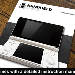 May include: A white handheld console with two black screens, buttons, and a directional pad, resting on a white and black printed sheet. The sheet has the words "HANDHELD CONSOLE" and "With your style." printed on it. The text "Comes with a detailed instruction manual" is at the bottom.