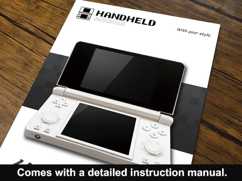 May include: A white handheld console with two black screens, buttons, and a directional pad, resting on a white and black printed sheet. The sheet has the words "HANDHELD CONSOLE" and "With your style." printed on it. The text "Comes with a detailed instruction manual" is at the bottom.