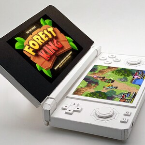 May include: A white handheld gaming console with a game screen displaying a colorful fantasy game. The top screen shows the text "FOREST KING" in a stylized font. The console's buttons and directional pad are white.