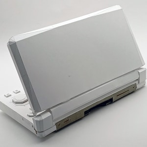 May include: A white handheld gaming console with a hinged design. The console is open, revealing the screen and control buttons. The device has a sleek, modern aesthetic with a minimalist design.