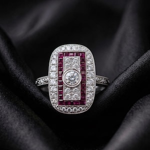 May include: A rectangular silver-toned ring featuring a central round brilliant-cut stone surrounded by a border of square-cut red rubies and a frame of small round white stones. The metal band displays intricate engraved scrollwork detailing.