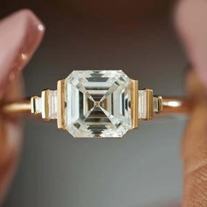 May include: A gold ring featuring a large, square-cut, clear gemstone as the centerpiece. The ring has smaller rectangular gemstones on each side of the main stone. The band is a thin, polished gold.