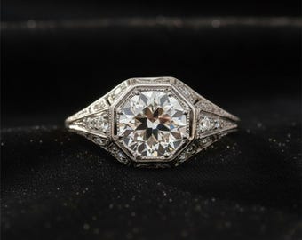 May include: A vintage-style engagement ring with a large, round, brilliant-cut diamond as the centerpiece. The diamond is set in an octagonal frame, with smaller diamonds and intricate filigree detailing on the band. The ring is made of white metal, likely platinum or white gold.