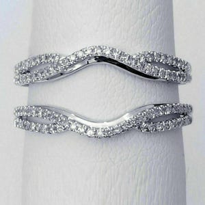 May include: Two silver-coloured, curved wedding bands. Each band is encrusted with small, clear gemstones. The bands are designed to interlock, creating a unique and elegant look. The bands are set against a white background.