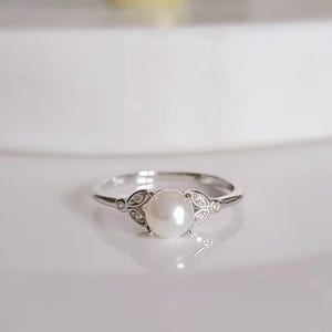 May include: A silver ring featuring a large, round, white pearl as the centerpiece. The ring has a delicate design with leaf-like accents and small, sparkling stones on either side of the pearl. The ring is set against a white background.