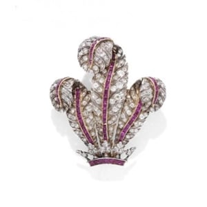 May include: A detailed brooch in the shape of a stylized feather design. The brooch features a silver-colored metal base, adorned with numerous small, clear gemstones and accented with vibrant, deep red gemstones. The design is symmetrical and elegant.