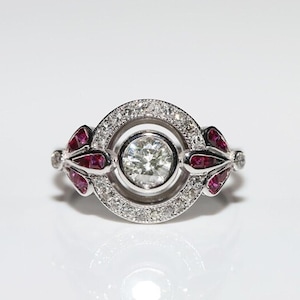 May include: A silver ring with a large round diamond surrounded by smaller diamonds. The ring has two ruby accents on either side of the diamond.