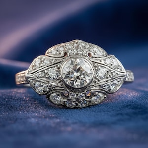 May include: A vintage-style ring with a large, round, brilliant-cut diamond center stone. The ring features intricate detailing with smaller diamonds set in a floral or art deco design, all set in a silver or platinum band. The ring is displayed on a blue velvet surface.