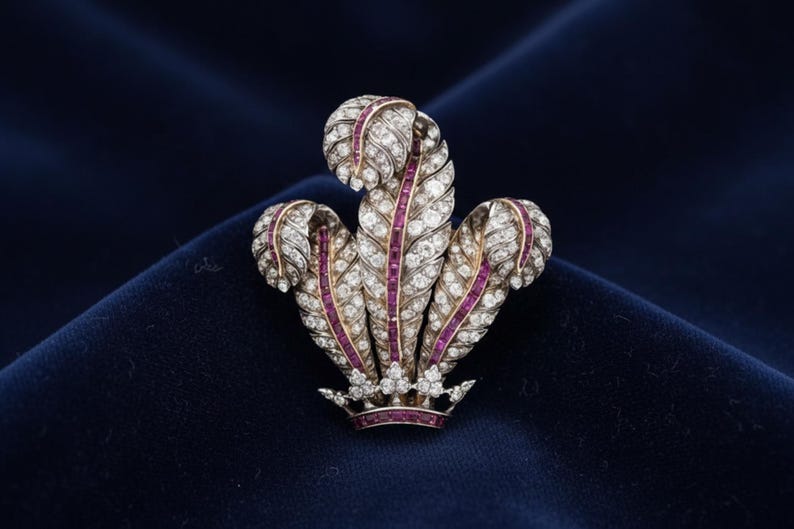 May include: A detailed brooch featuring a design of three stylized feathers, adorned with numerous small, sparkling diamonds. The feathers have a central band of red gemstones, and the base is a small crown. The brooch is set against a dark blue background.