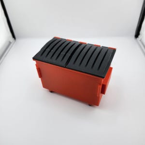 Puede incluir: Un pequeño contenedor de basura rojo con tapa negra. La tapa tiene líneas curvas y paralelas. El contenedor es de forma rectangular y tiene pequeñas ruedas.