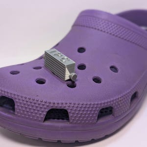 May include: A purple clog with a silver charm attached to the strap. The charm is a small, rectangular object with the word "LEGO" printed on it.