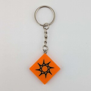 Pode incluir: Chaveiro laranja em forma de diamante com um desenho de explos&atilde;o solar preto. O chaveiro tem um anel e uma corrente prateados. O desenho est&aacute; centrado no fundo laranja. Um acess&oacute;rio simples, mas atraente.