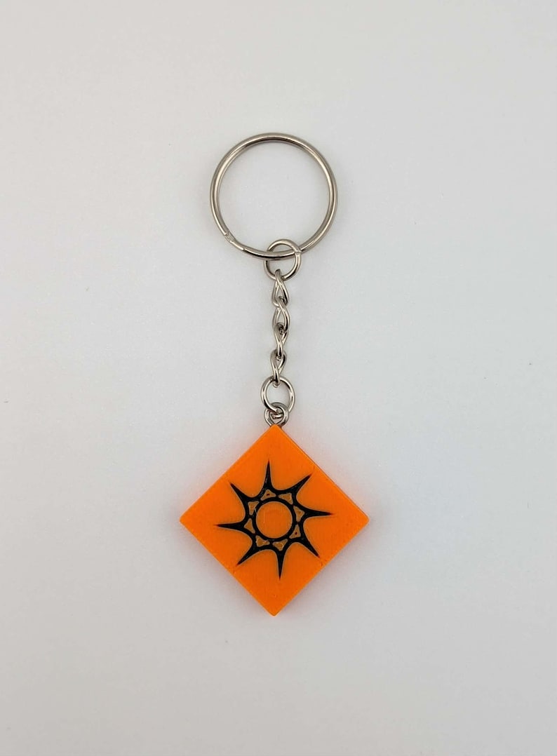 Pode incluir: Chaveiro laranja em forma de diamante com um desenho de explos&atilde;o solar preto. O chaveiro tem um anel e uma corrente prateados. O desenho est&aacute; centrado no fundo laranja. Um acess&oacute;rio simples, mas atraente.
