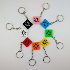 May include: A collection of colorful square keychains. Each keychain features a different color square with a sunburst design in the center. The keychains are attached to silver key rings and chains. Colors include black, white, pink, blue, green, yellow, orange, and red.