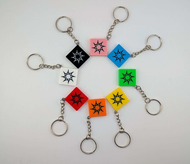 Pode incluir: Uma cole&ccedil;&atilde;o de chaveiros quadrados coloridos. Cada chaveiro apresenta um quadrado de cor diferente com um desenho de sol no centro. Os chaveiros s&atilde;o presos a argolas e correntes prateadas. Cores: preto, branco, rosa, azul, verde, amarelo, laranja e vermelho.