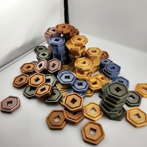 Può includere: Una collezione di gettoni esagonali in vari colori, tra cui bronzo, oro, blu e verde. Ogni gettone ha un ritaglio quadrato al centro. I gettoni sono impilati e sparsi, suggerendo un gioco o un componente artigianale.