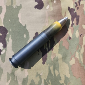 May include: A dark blue, cylindrical object with a silver tip and a yellow band near the top. The object is lying on a camouflage pattern fabric. The object has a black base.