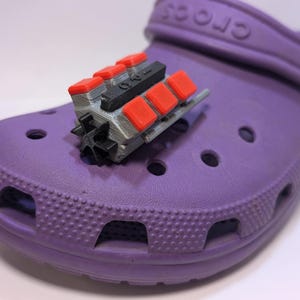 Może przedstawiać: Fioletowy but Croc z zawieszką w kształcie silnika, wydrukowaną w 3D w kolorze czerwonym i szarym, przymocowaną do paska.