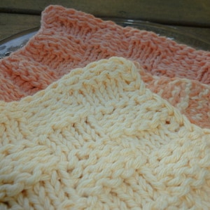 May include: Two knitted dishcloths, one in a pale yellow and one in a peach color. The dishcloths are made with a simple knit stitch and have a textured appearance.