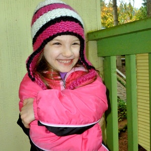 May include: A young girl wearing a pink and white striped knit hat with black accents and a pink jacket with black trim. She is smiling and has her arms crossed.