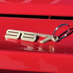 May include: A silver metal keychain with the number "987" attached to a silver ring. The keychain is on a red surface.