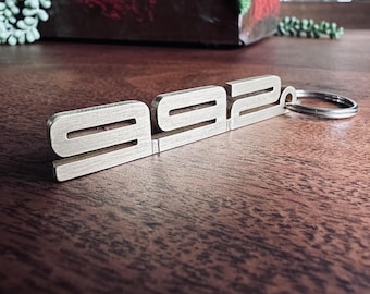 Puede incluir: Un llavero de metal plateado con el número "992" grabado en él. El llavero está unido a un anillo de metal plateado.