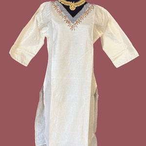 May include: A long-sleeved, off-white tunic featuring a V-neckline with intricate pink and green floral embroidery along the collar. The garment has side slits and is displayed with a gold-toned necklace featuring a central circular pendant.