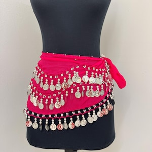 May include: A hot pink belly dance hip scarf with silver coin embellishments. The scarf is tied around a black mannequin. The coins are attached in rows and hang down, creating a decorative fringe.