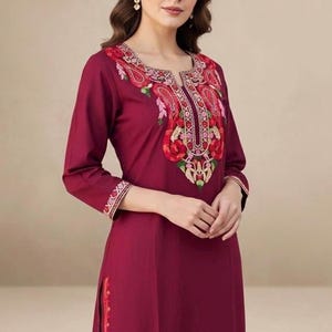Peut inclure: Robe tunique bordeaux avec broderies florales complexes autour de l'encolure et des manches. La robe présente un design long et fluide avec une fente latérale et une bordure détaillée. La broderie comprend des motifs floraux rouges, roses et verts.