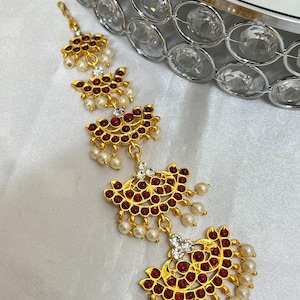 May include: A gold-toned headpiece with a series of fan-shaped pendants. Each pendant is adorned with small red gemstones and faux pearls. Clear crystals are set between the pendants. The headpiece is designed for formal occasions.