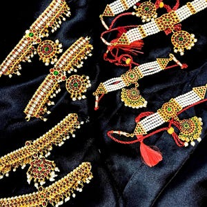 May include: An assortment of gold-toned jewelry sets, including necklaces and earrings, are displayed on a black surface. The jewelry features red and green gemstones, white beads, and intricate designs. Some sets have red tassels.