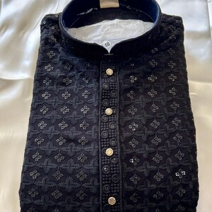 May include: A black embroidered kurta with a stand-up collar and button closure. The kurta has a geometric pattern and is decorated with gold buttons.