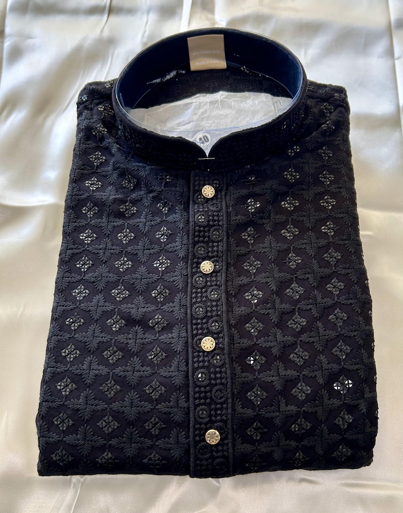 May include: A black embroidered kurta with a stand-up collar and button closure. The kurta has a geometric pattern and is decorated with gold buttons.