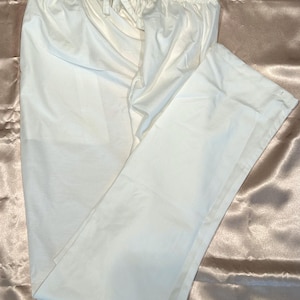 May include: White cotton pants with a drawstring waist. The pants are folded and laid flat on a light brown satin fabric.
