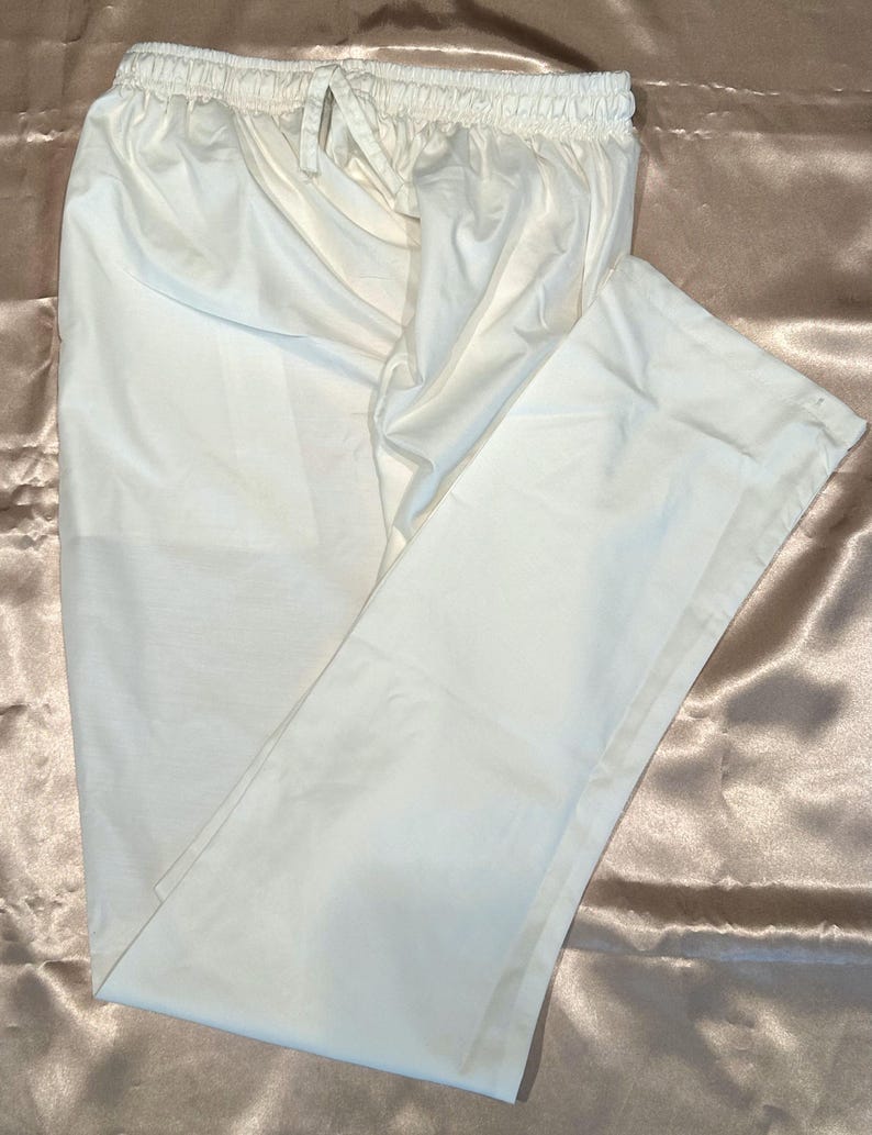May include: White cotton pants with a drawstring waist. The pants are folded and laid flat on a light brown satin fabric.