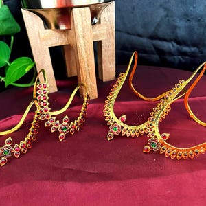 以下が含まれることがあります： 赤と緑の宝石がアクセントになった金色のヘッドピース。ジュエリーは、複雑な花柄と幾何学模様が特徴です。作品は、木製のスタンドと背景の緑とともに、バーガンディ色の表面に展示されています。