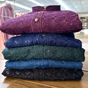 May include: A stack of five embroidered shirts in different colors, including purple, blue, green, and black. The shirts have a geometric pattern and are folded neatly.